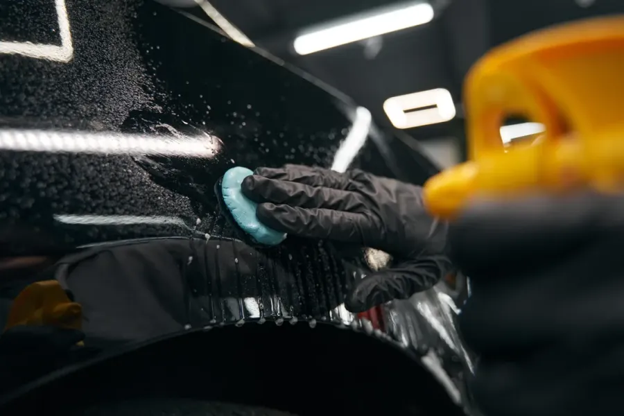 Behandschuhte Hand zieht blaue Reinigungsknete über eine nasse schwarze Lackfläche